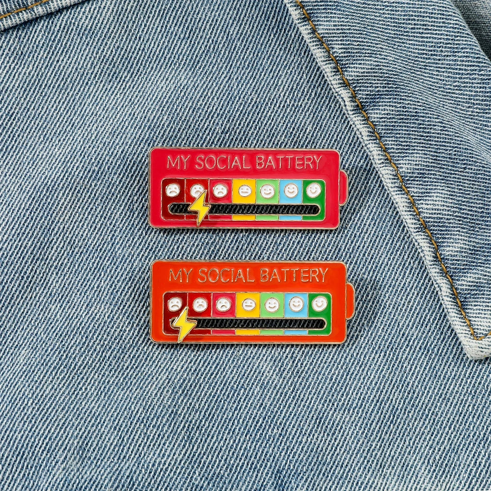 Two red mood battery pins with smiley faces and lightning bolts pinned onto a denim fabric.