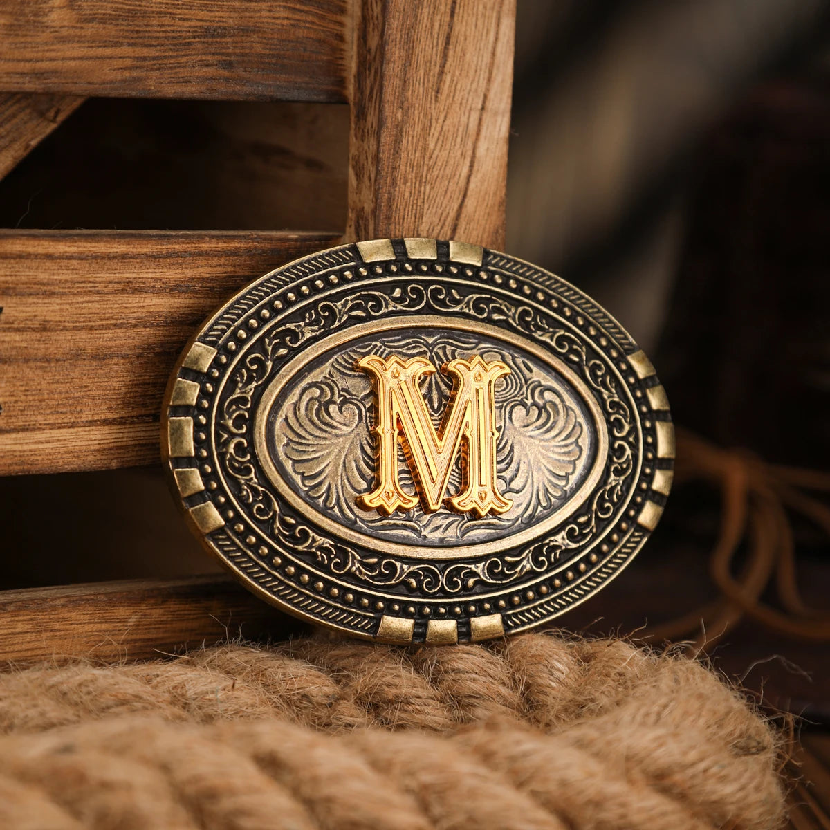 A vintage western belt buckle with a gold-toned 'M' design set in an ornate silver-toned circle, resting on a brown wooden su