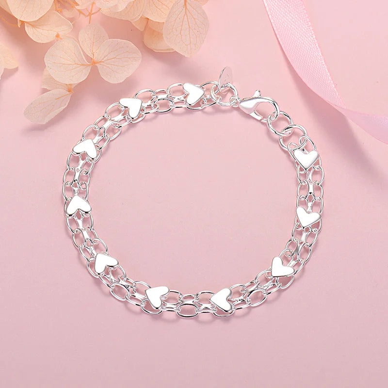 A silver heart charm bracelet made of 925 sterling silver, displayed on a pink background.