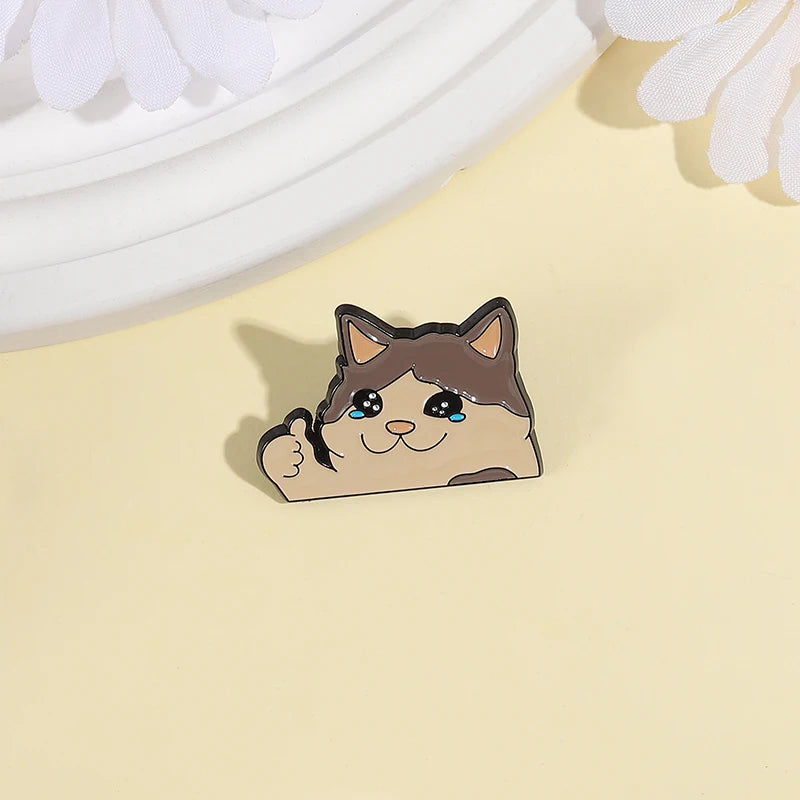 A cute enamel pin of a cat with teary eyes and a happy expression resting on a light yellow background.