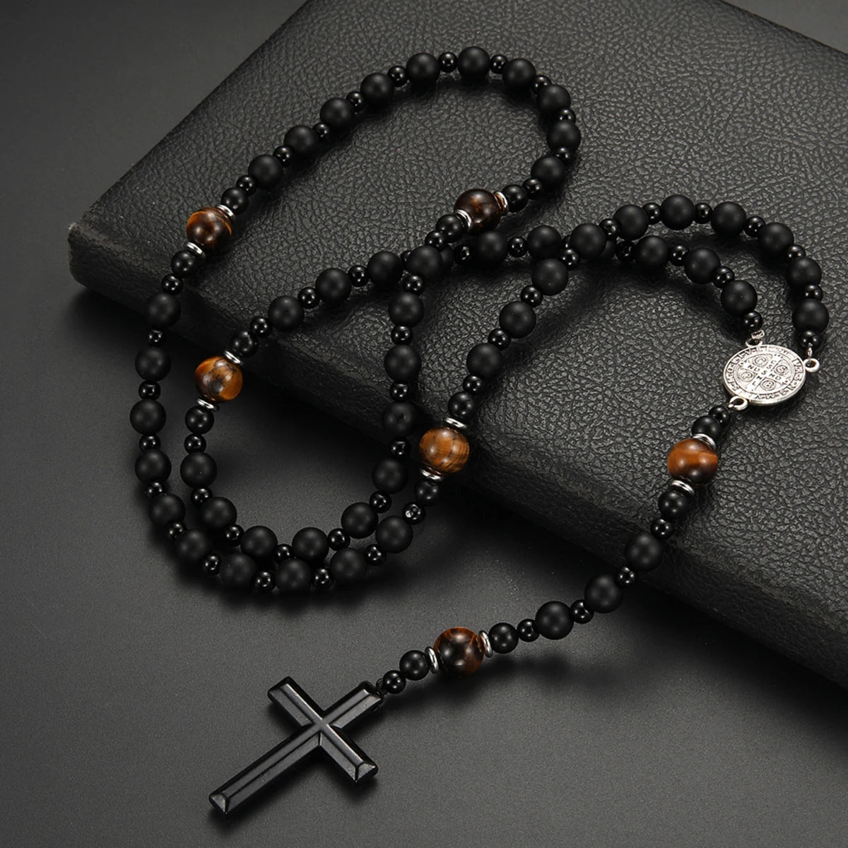 A black obsidian and tiger's eye beaded cross pendant necklace for men displayed on a dark surface.