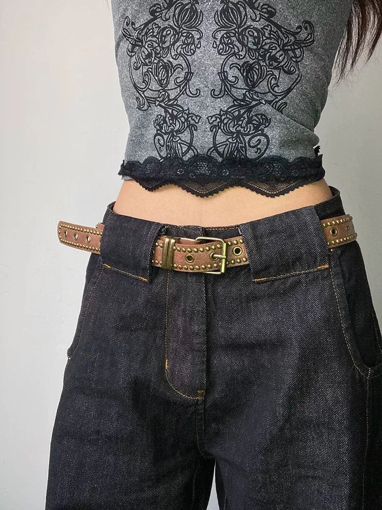 A close-up of dark denim jeans and a gray sheer top with black lace trim, featuring a brown leather belt with bronze rivets.