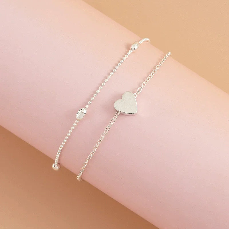 Un anklet argenté à perles délicat avec un charm en forme de cœur sur le poignet d'une personne.
