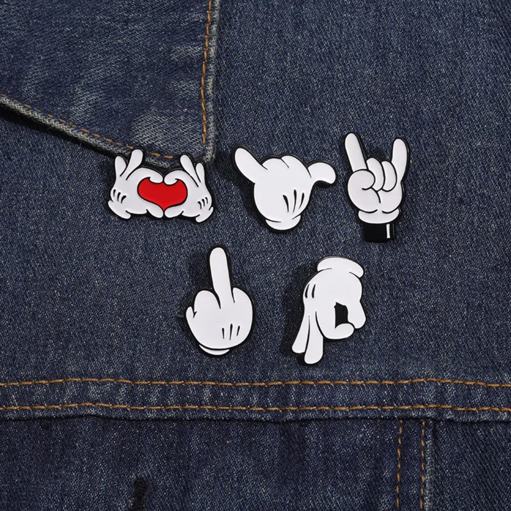 A close-up of a dark denim fabric featuring white enamel pins depicting various finger gestures and a heart.