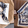 A close-up of a patterned silk scarf tied in a bow, displayed against a neutral beige jacket and a folded scarf.