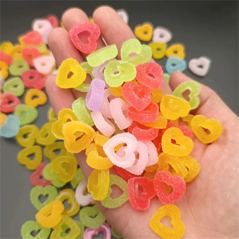 A hand holds a collection of colorful heart-shaped resin nail art charms in various shades of pink, yellow, green, and orange
