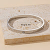 A silver stainless steel bangle bracelet is displayed on a light beige textured surface.