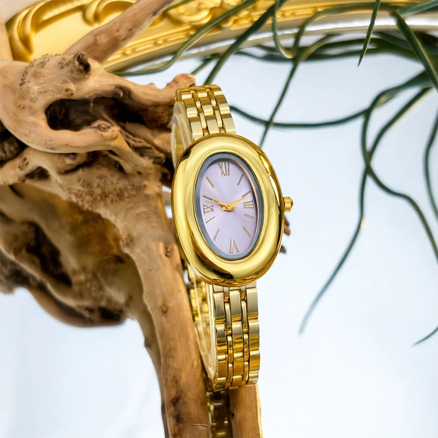 Un reloj vintage con caja dorada y esfera rosa claro, exhibido en una rama de madera.