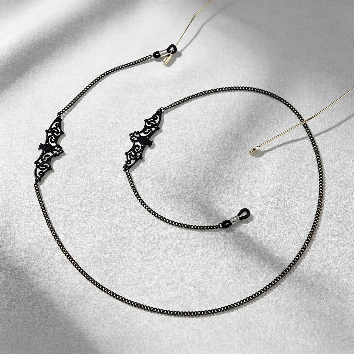 A delicate black metal link strap for glasses and sunglasses is displayed on a light gray fabric.