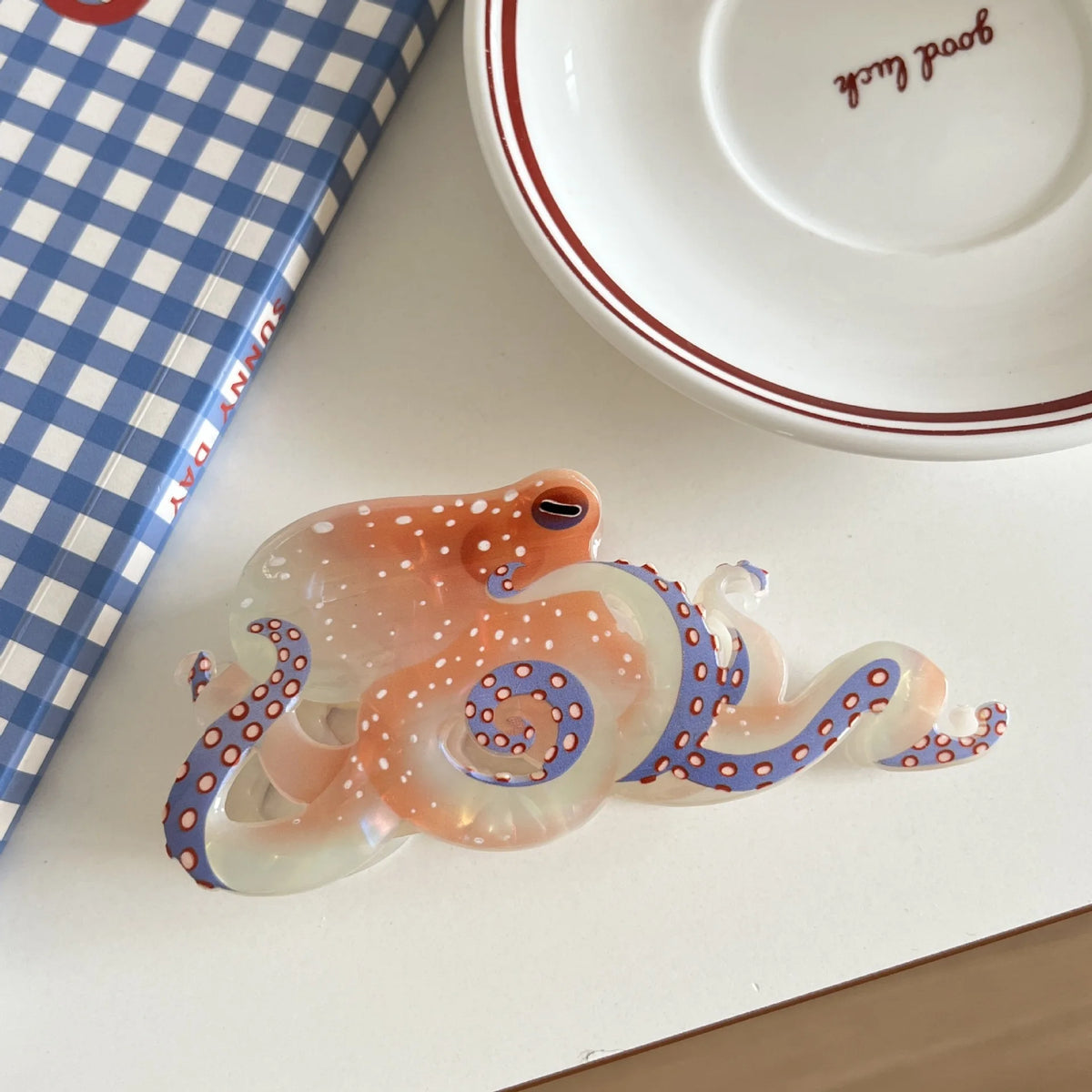 Pince à cheveux en forme d'octopus, orange et blanc translucide avec des pois bleus, posée sur une surface blanche.