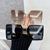 A hand holds a pair of elegant square sunglasses, featuring a clear pink-toned frame and dark lenses.