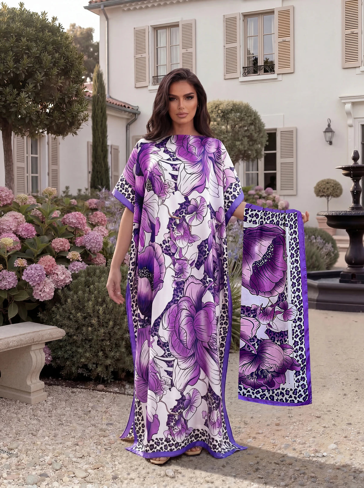A woman models a purple and white tropical floral print abaya scarf outdoors in front of a white building.