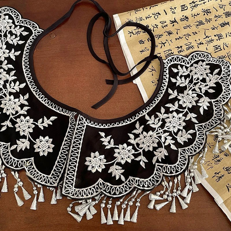 Chemise à col de dentelle noire avec broderies blanches et franges sur une surface en bois brun.