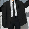 A person wearing a black suit jacket, white shirt, and black tie standing in front of a light-colored surface.