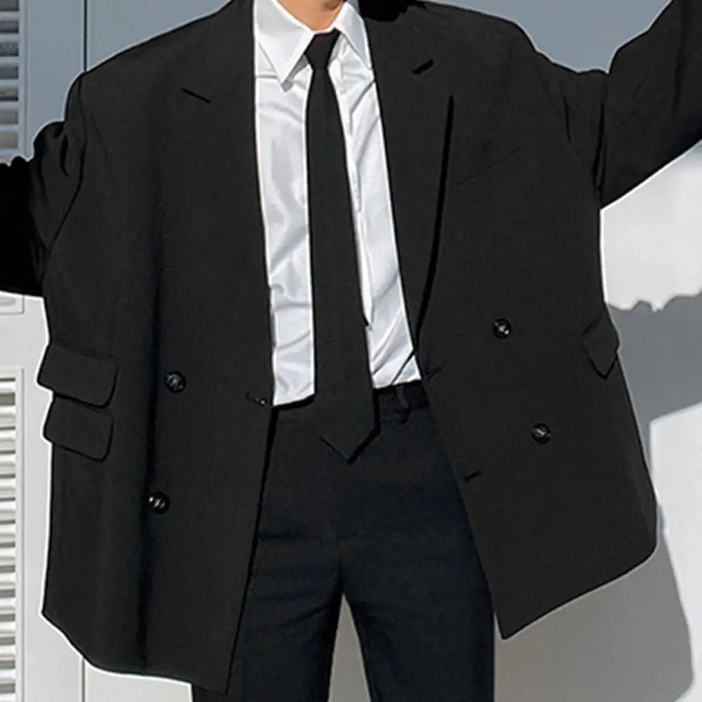 A person wearing a black suit jacket, white shirt, and black tie standing in front of a light-colored surface.