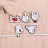 Enamel pin brooch featuring cartoon finger gestures including a heart and a peace sign on a pink fabric background