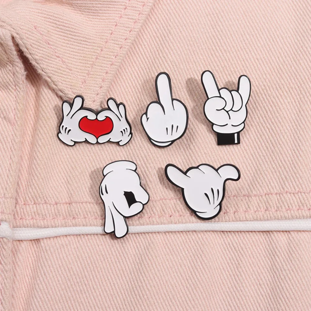 Enamel pin brooch featuring cartoon finger gestures including a heart and a peace sign on a pink fabric background