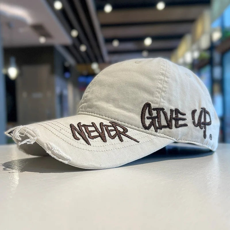 A light beige baseball cap with embroidered text that reads NEVER GIVE UP resting on a smooth surface.