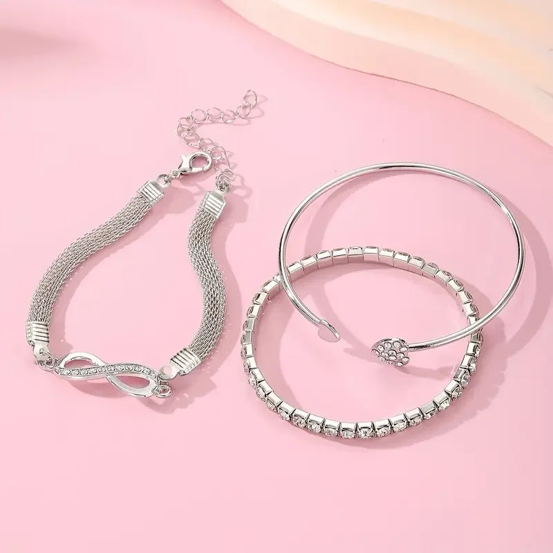 A set of three silver heart charm bracelets with delicate chains and crystals displayed on a pink background.
