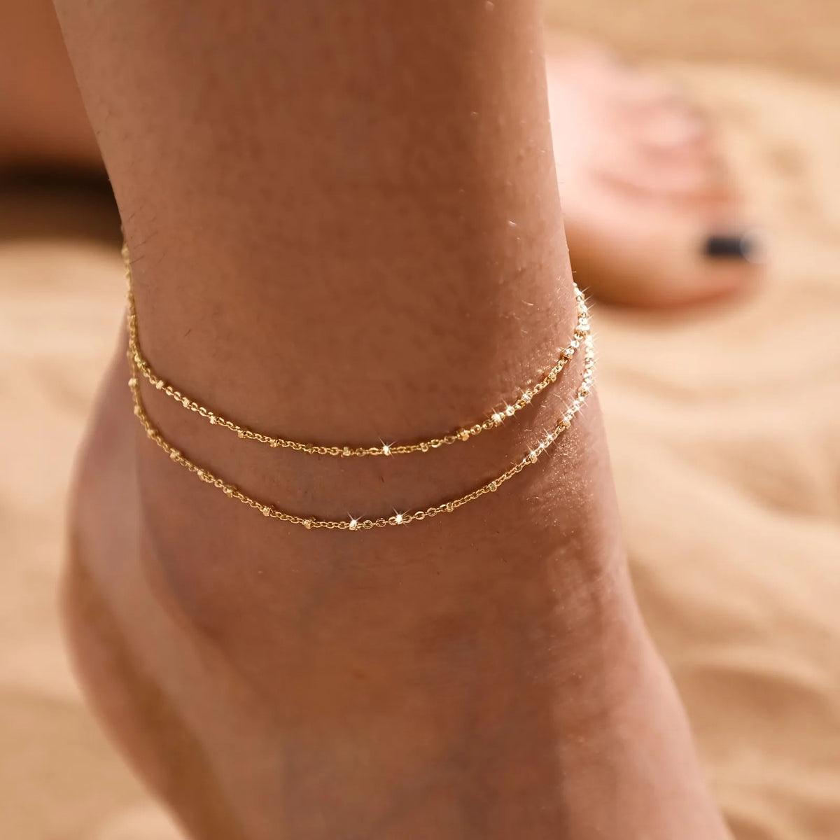 Primer plano de un tobillo con dos delicadas pulseras de acero inoxidable doradas.
