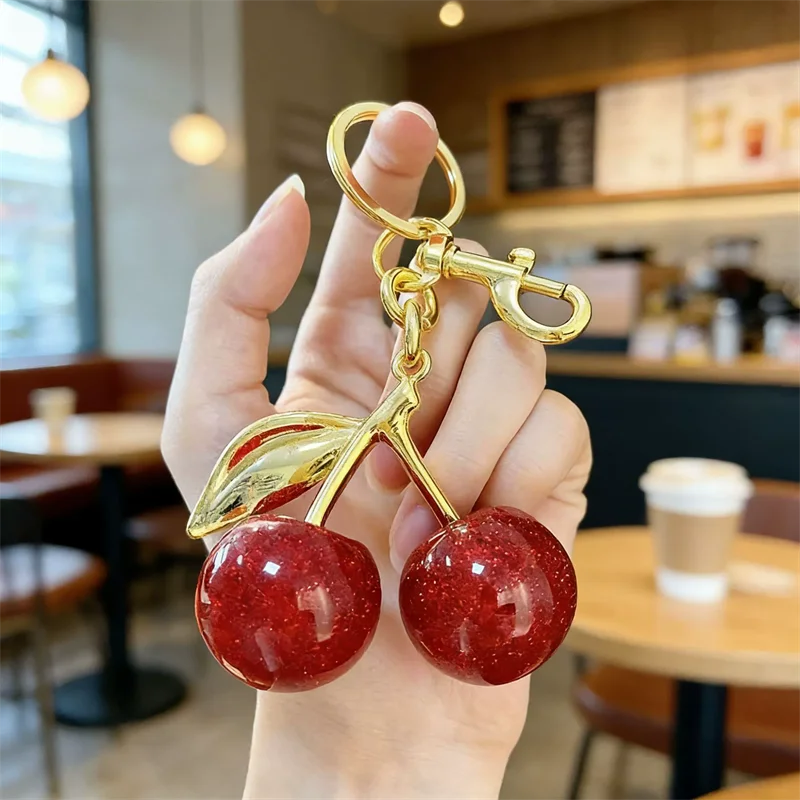 A hand holds a gold-colored key chain decoration featuring two shiny red cherry beads on stems.