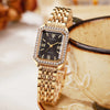 Reloj de cuarzo dorado con esfera cuadrada, dial negro y detalles de strass, sobre un cuenco de cerámica.