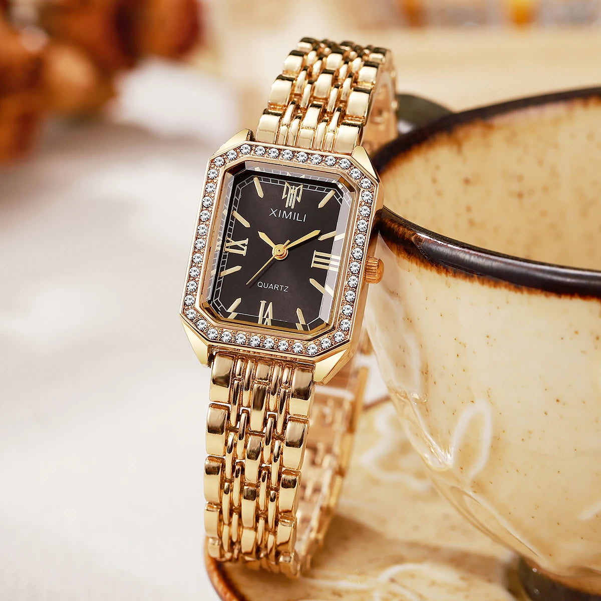 Reloj de cuarzo dorado con esfera cuadrada, dial negro y detalles de strass, sobre un cuenco de cerámica.