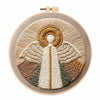 A circular embroidery kit featuring a beige and gold angel design on a natural linen background.