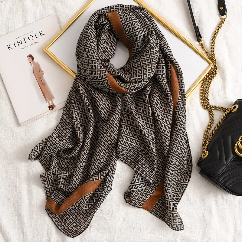 Dunkel gemusterter Seid-Pashmina-Schal auf weißer Fläche neben einem goldenen Rahmen und einer schwarzen Ledertasche.