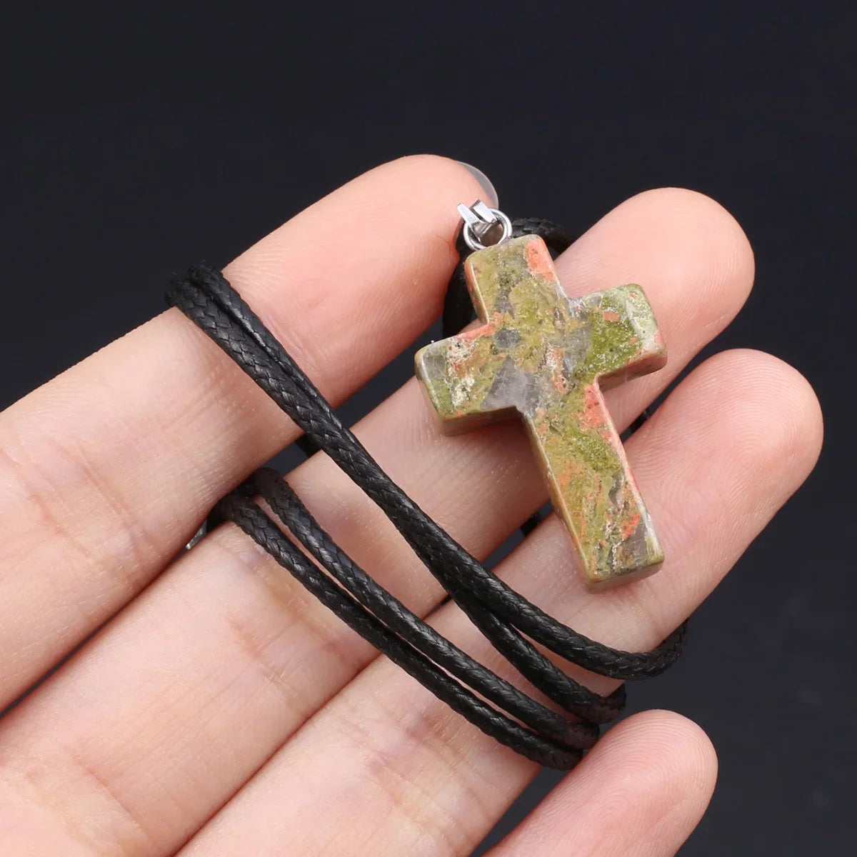 Eine Hand hält eine Tigerauge-Agat-Kreuz-Anhänger-Kette mit einem grünlich-gelben Stein und einem schwarzen Band.
