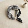 A black and white checkered silk scarf tied with a satin bow and a pearl magnetic buckle on a beige surface.