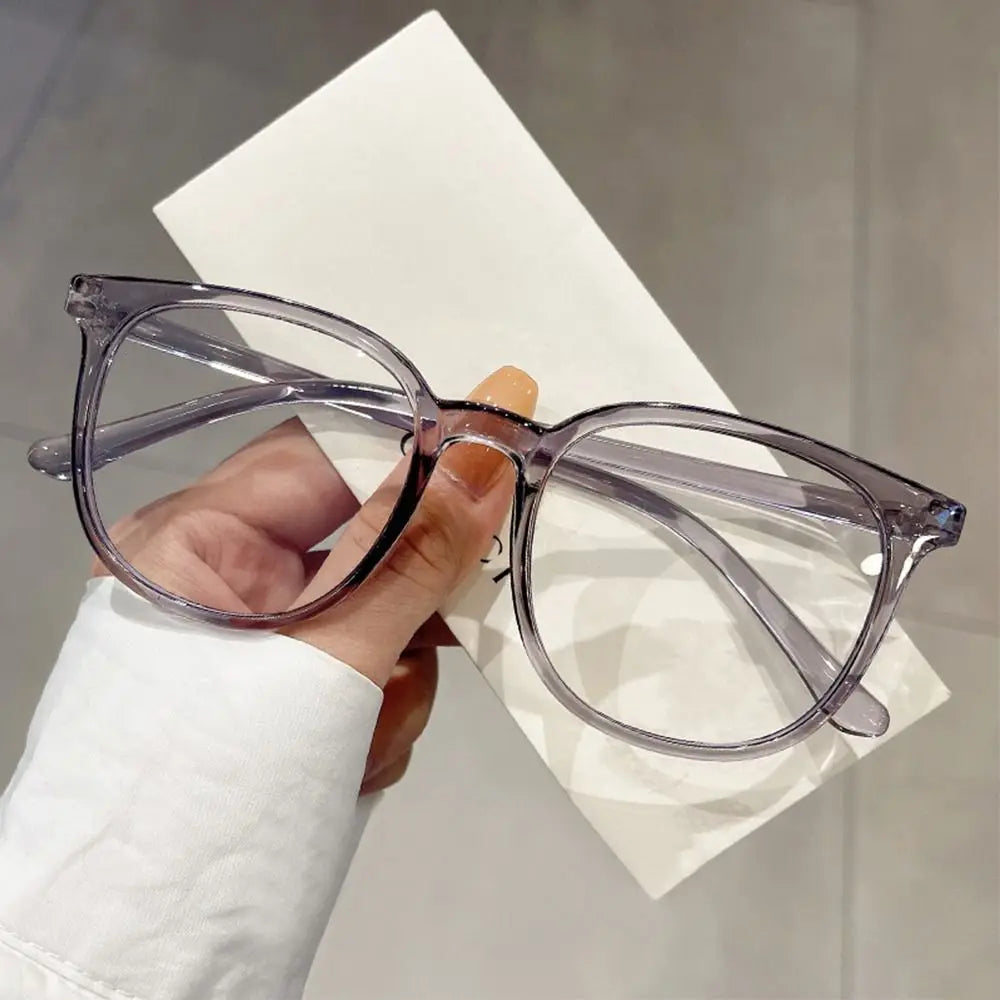 A hand holds a pair of clear, large square anti-blue light spectacles resting on a white card.