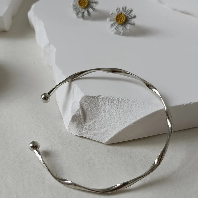 Silver twisted wave cuff bracelets displayed on a white stone next to small daisy-like flowers.