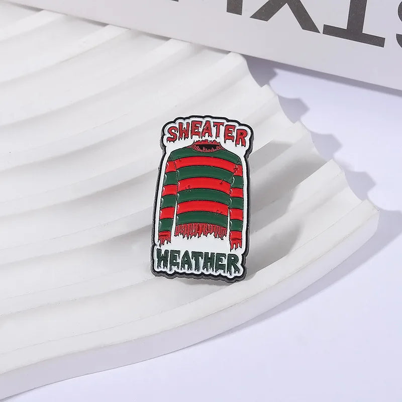 An enamel lapel pin featuring a striped green and red sweater design with the text SWEATER and WEATHER.