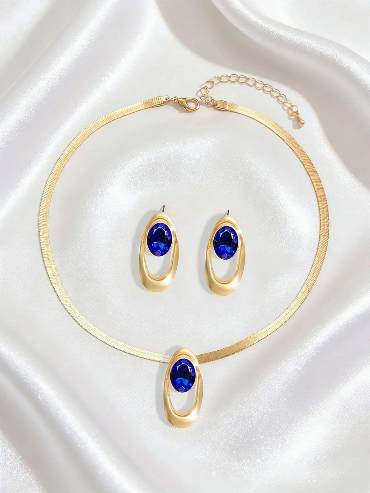 Gold necklace set featuring three oval blue gemstone rings and a matching bracelet displayed on silk fabric.
