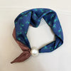 A navy blue silk scarf with a green deer print tied in a knot and secured with a pearl magnetic clasp.