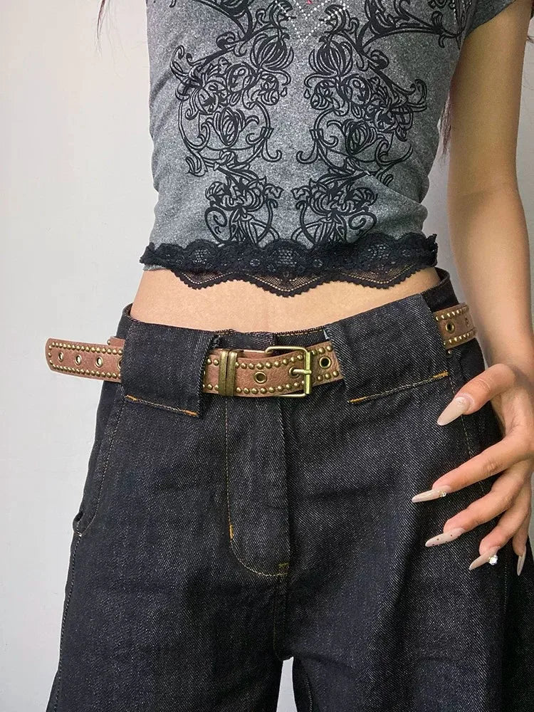 A close-up of dark denim pants and a gray floral top paired with a brown leather belt featuring bronze rivets.