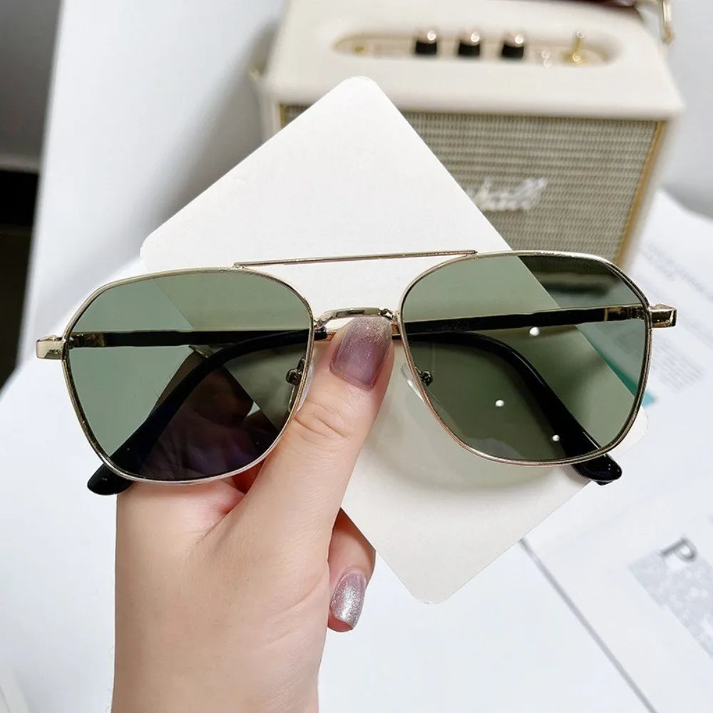 A hand holds a pair of classic metal frame square sunglasses with olive green lenses against a white background.