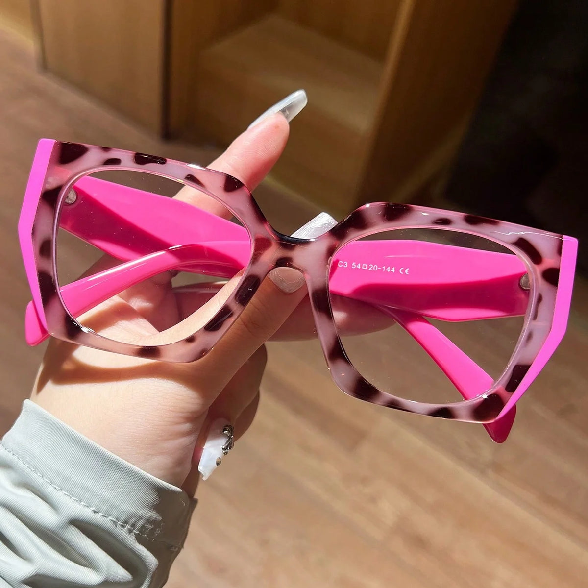 A hand holds a pair of pink leopard print fashion glasses with large frames.