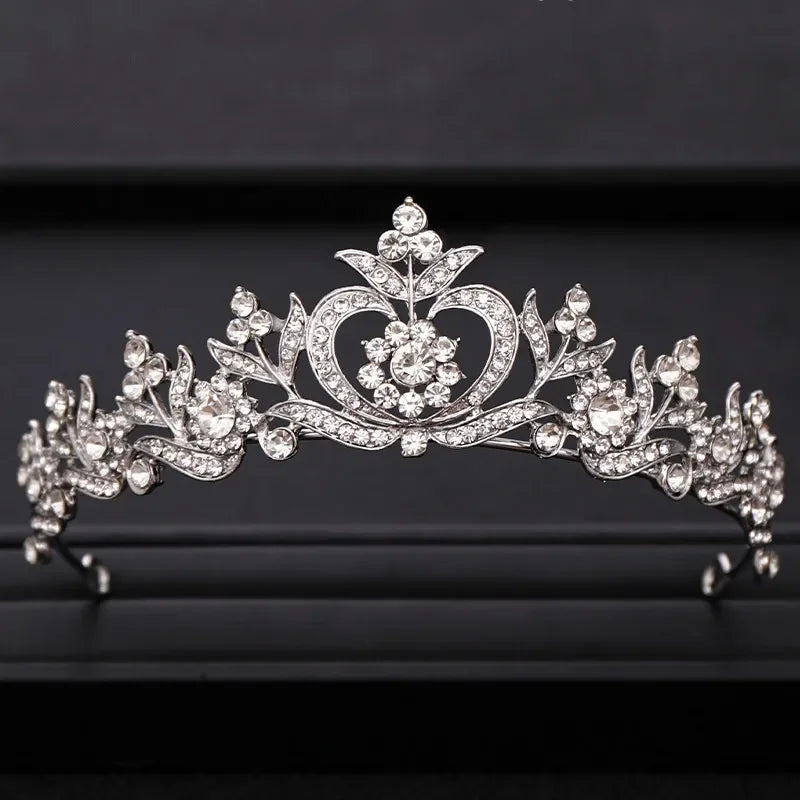 A sparkling crystal bridal crown with intricate floral and heart designs displayed on a dark background.