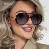 Eine Frau modelliert retroverlaufene Oversize-Sonnenbrillen mit blondem Haar und einem neutralen Blazer.