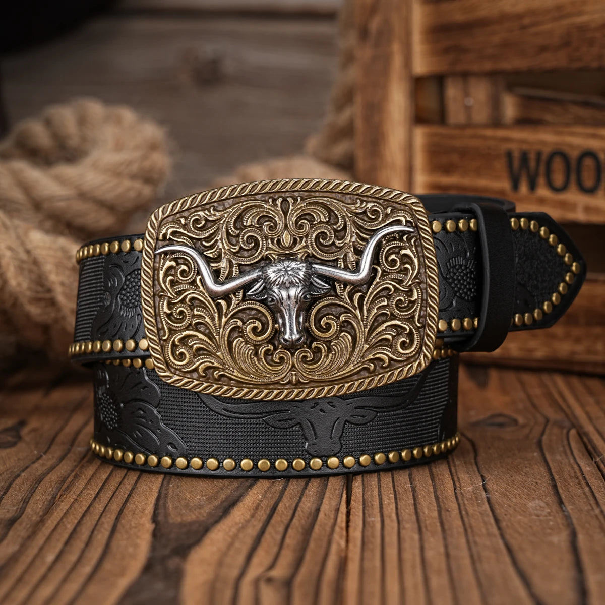 A black PU leather belt with a large, ornate gold-colored floral engraved buckle resting on a wooden surface.
