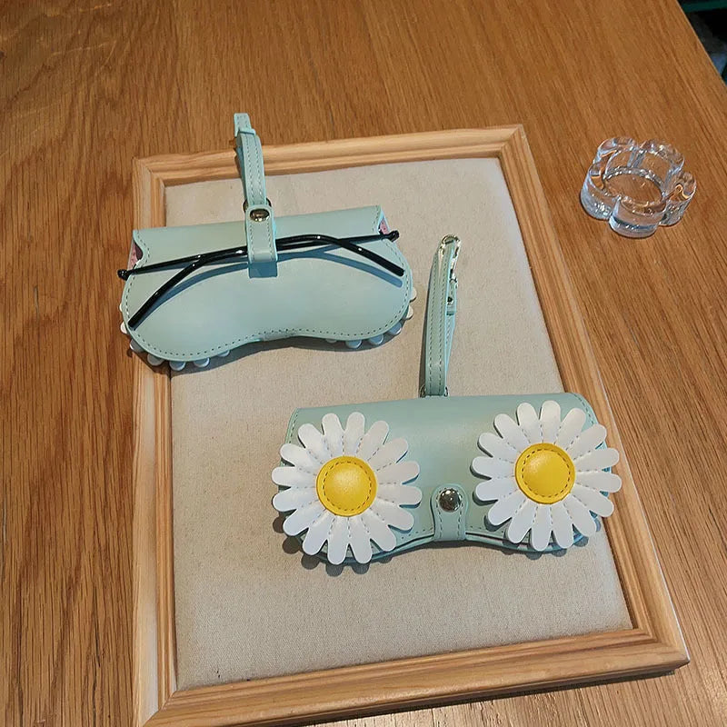 A light blue portable eyewear storage pouch with a black strap is displayed in a wooden frame on a table.