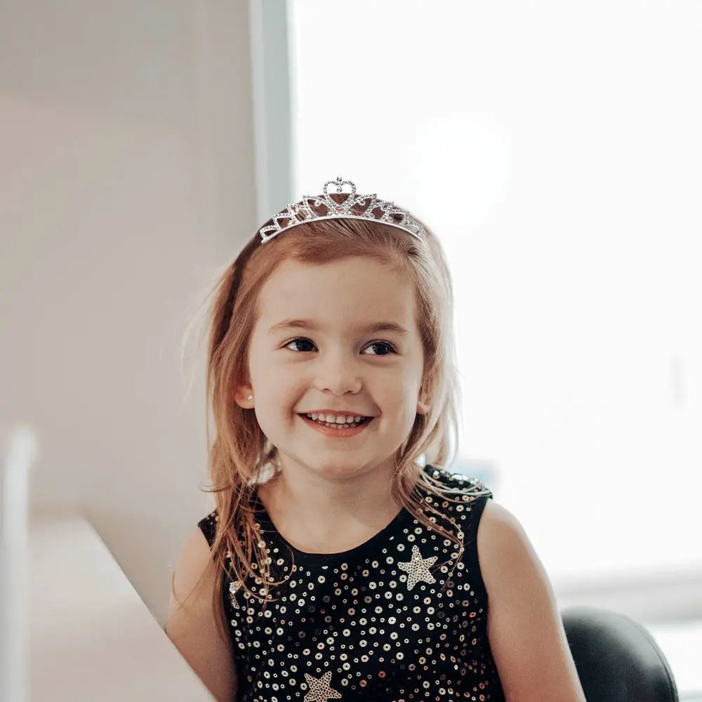 A young girl wearing a crystal crown smiling while sitting indoors.