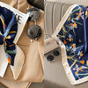 A navy blue silk scarf with a floral print featuring orange and blue flowers is displayed on a beige fabric.