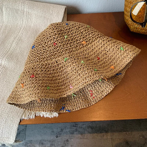 A close-up of a hand-crocheted Panama straw sun hat in natural beige with colorful pom-poms, resting on a brown wooden surfac