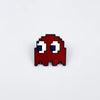 A red enamel pin shaped like Pac-Man's face with blue eyes and a white mouth.