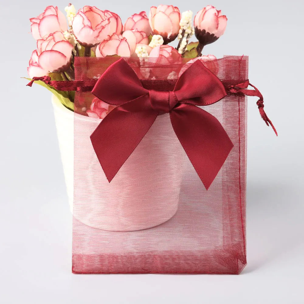A pink and white floral arrangement is presented in a deep red organza drawstring gift bag.