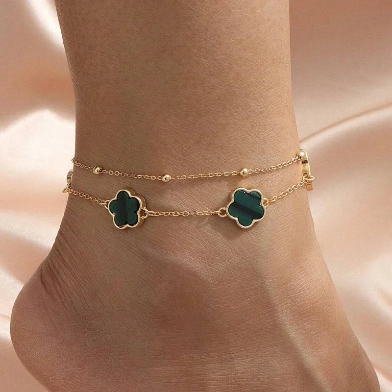 Gold chain anklet with five-leaf emerald green charms worn on a slender ankle.