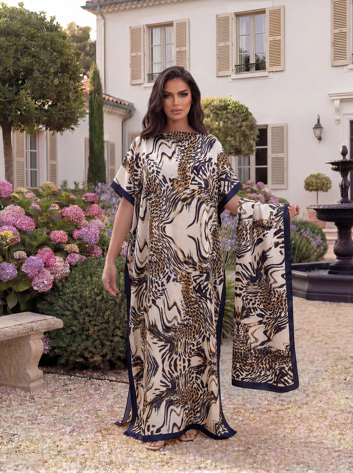 Eine Frau modelliert ein beige-braunes Leopardenmuster-Satinstoff-Abaya-Schalset in einer Gartenumgebung.